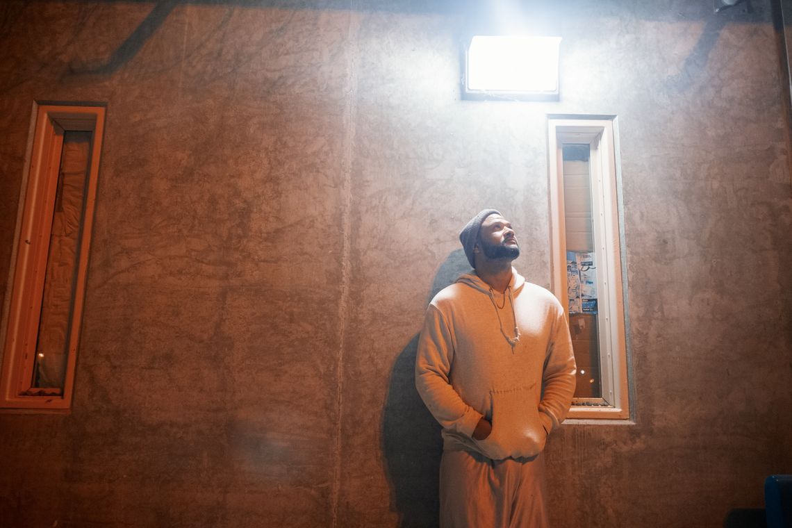 A photo shows a man with medium-toned skin and a beard, wearing a beanie and a light-colored hoodie sweatshirt, leaning against a wall with a light shining above him. 