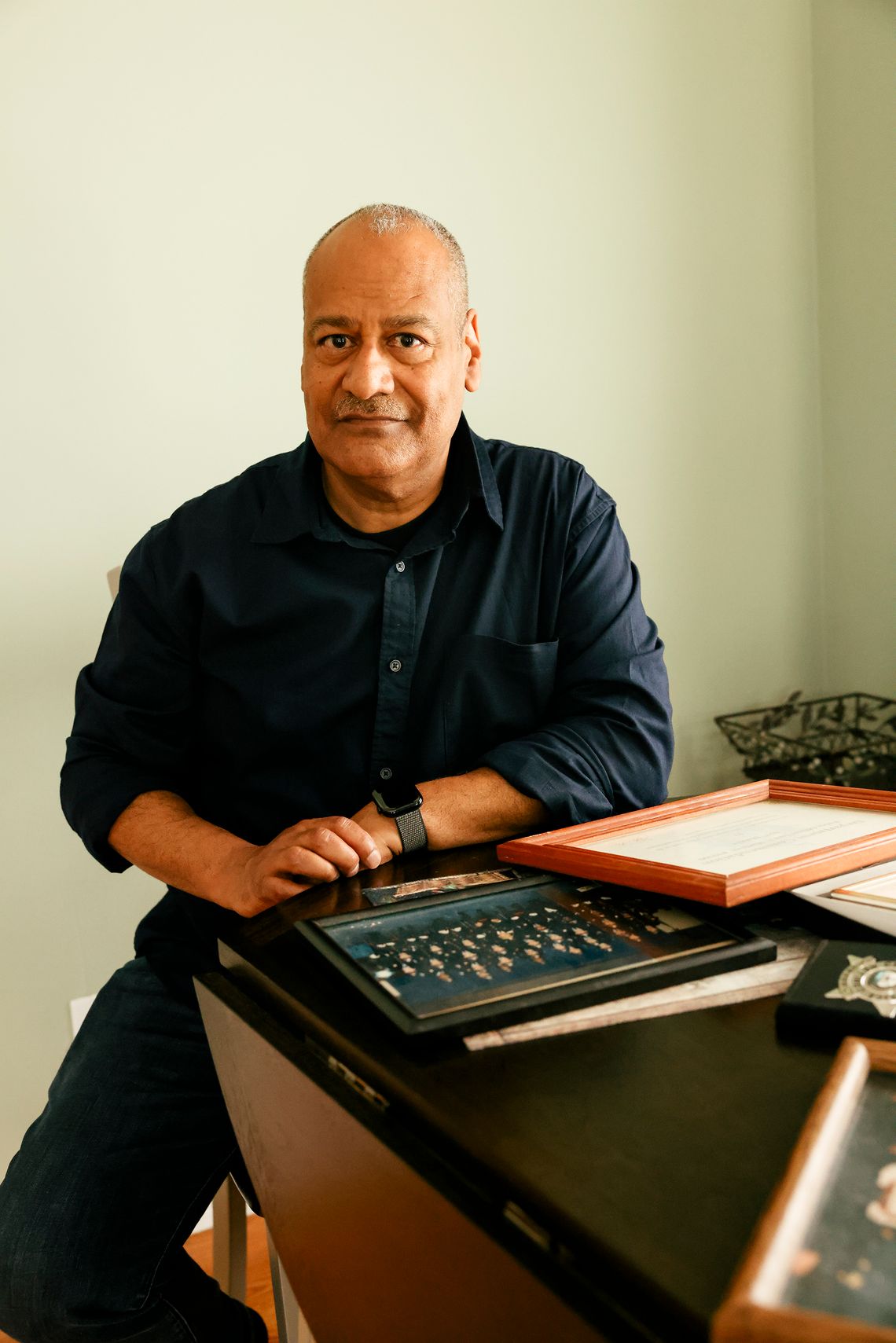 A photo shows Louis Martinez, a man with medium-toned skin, sitting at a table that has photo frames and certificate frames on it.