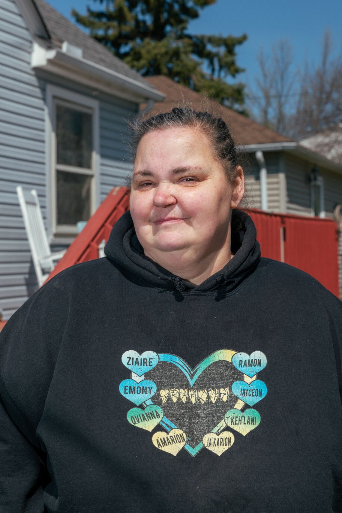 A vertical photo shows a White woman with brown hair tied back.She is wearing a black hoodie sweatshirt with hearts on the front. Behind her, part of a house is visible. 
