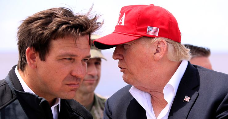 A White man in a black jacket leans his head forward as another White man, who is wearing a red cap and suit, talks to him. 