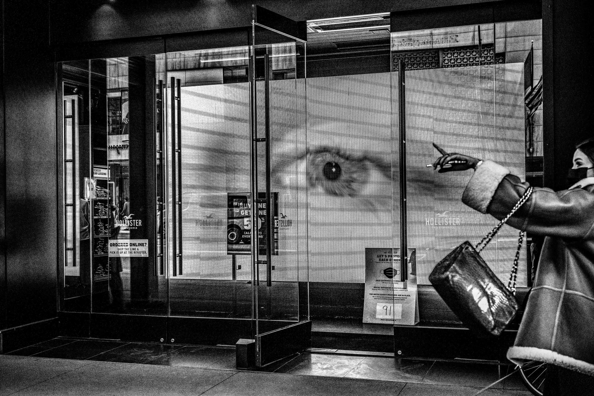 A black and white photograph shows a large electronic billboard with a closeup of an eye at the front of a store in New York.  