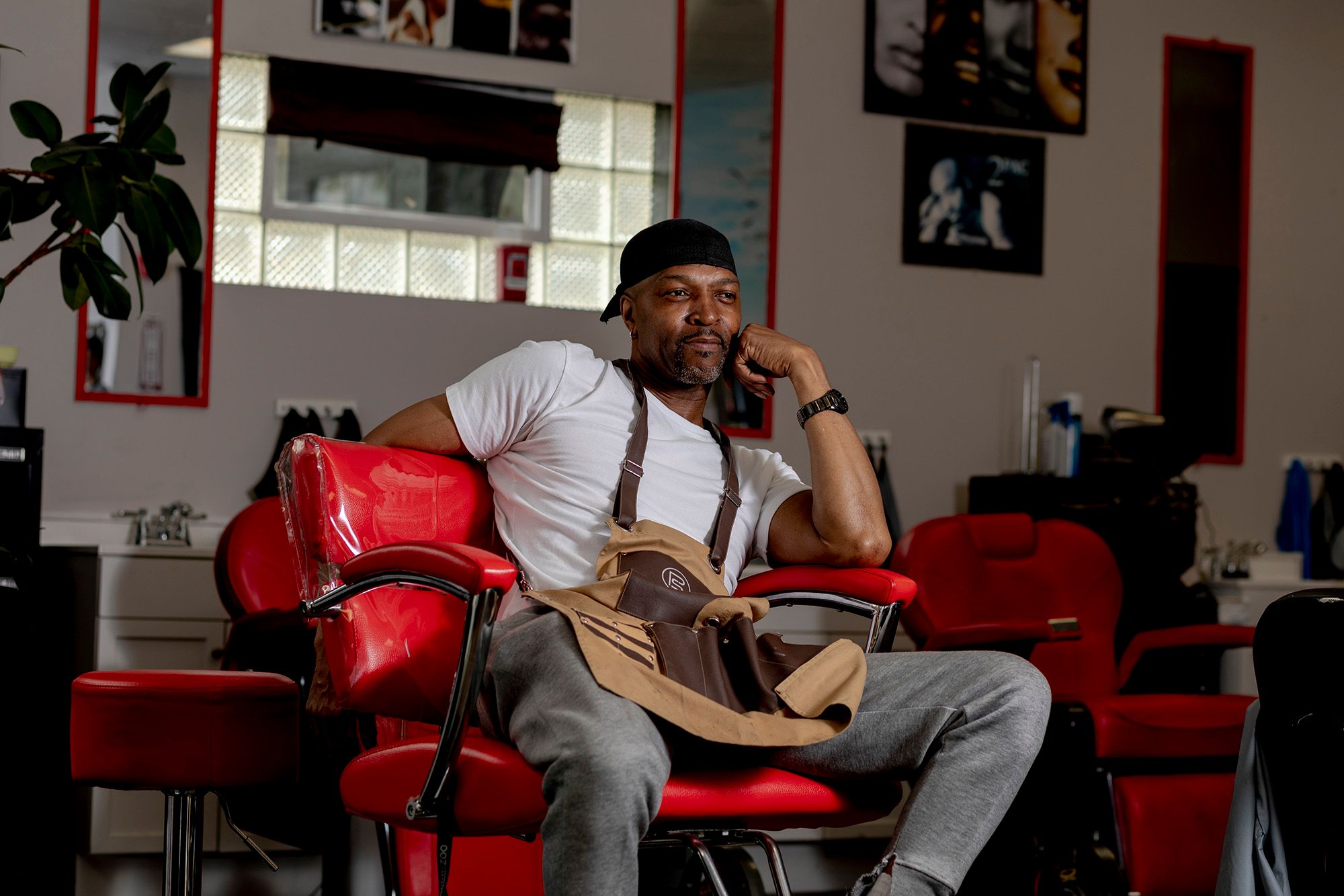 A Black man, in a white T-shirt, brown apron, and gray pants, sits on a red barber chair.