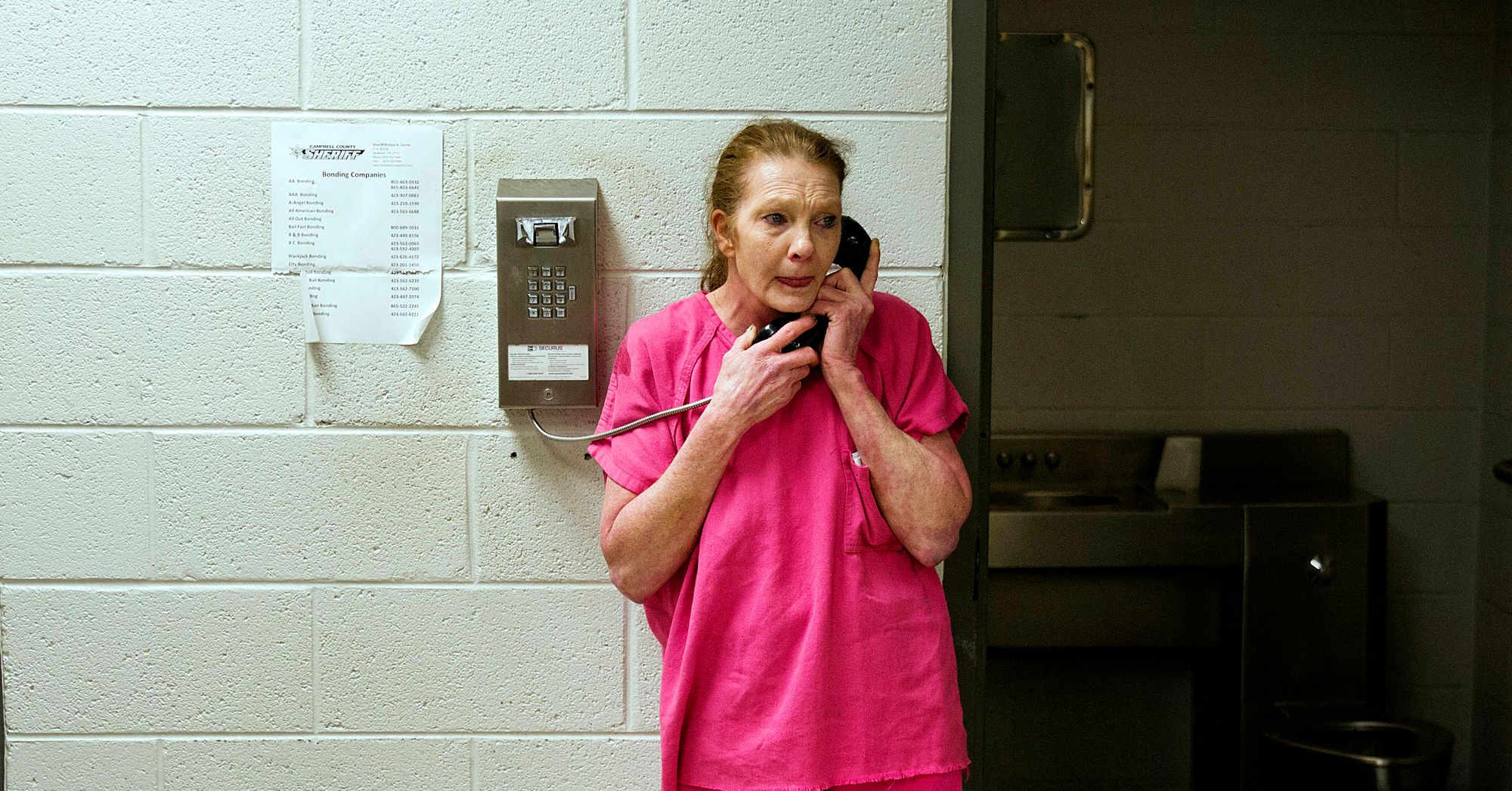 A White woman in a pink jail uniform stands against a brick wall as she makes a phone call. 