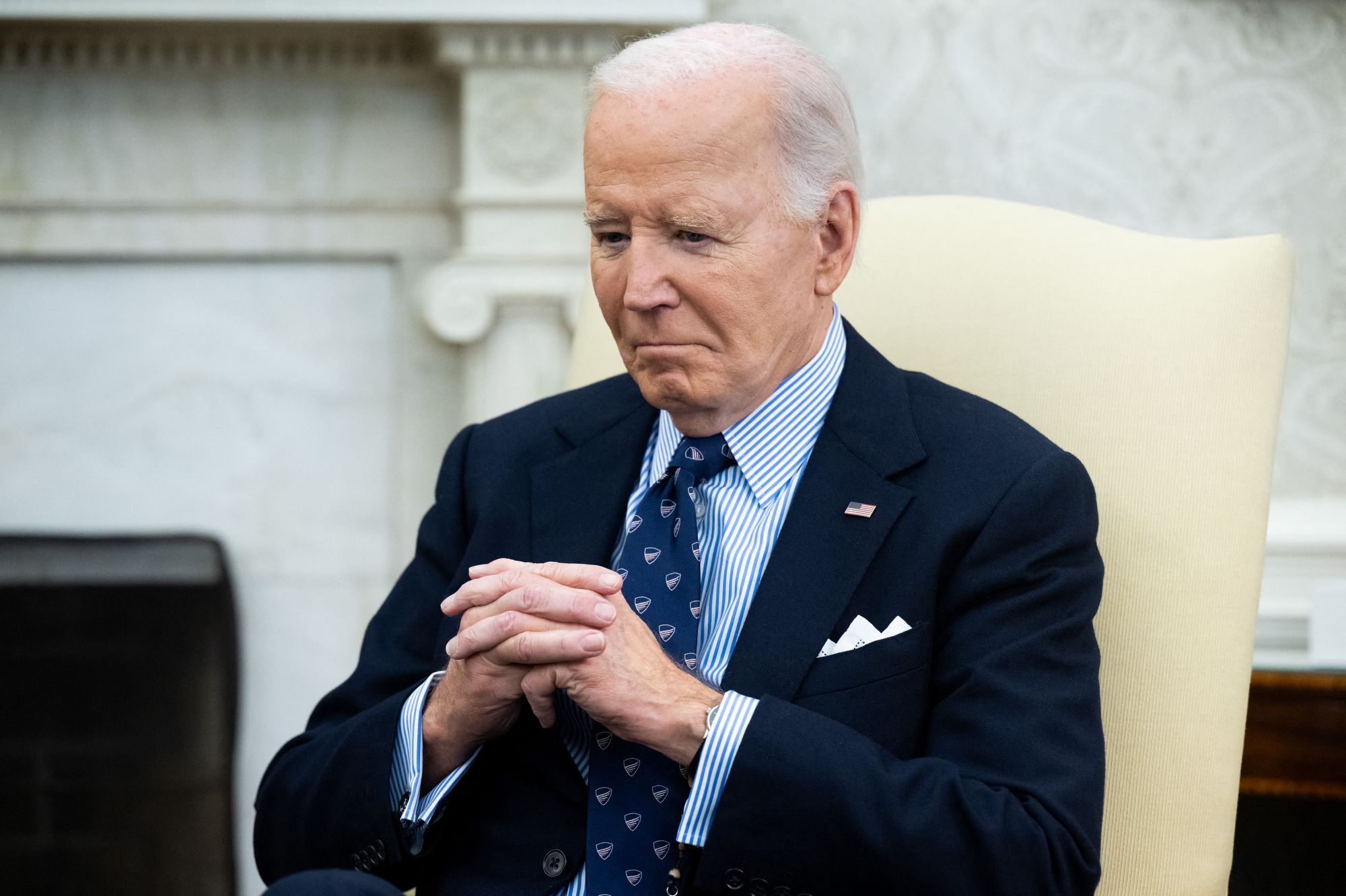 U.S. President Joe Biden sits in a white chair in a navy suit with his hands clasped in front of him.