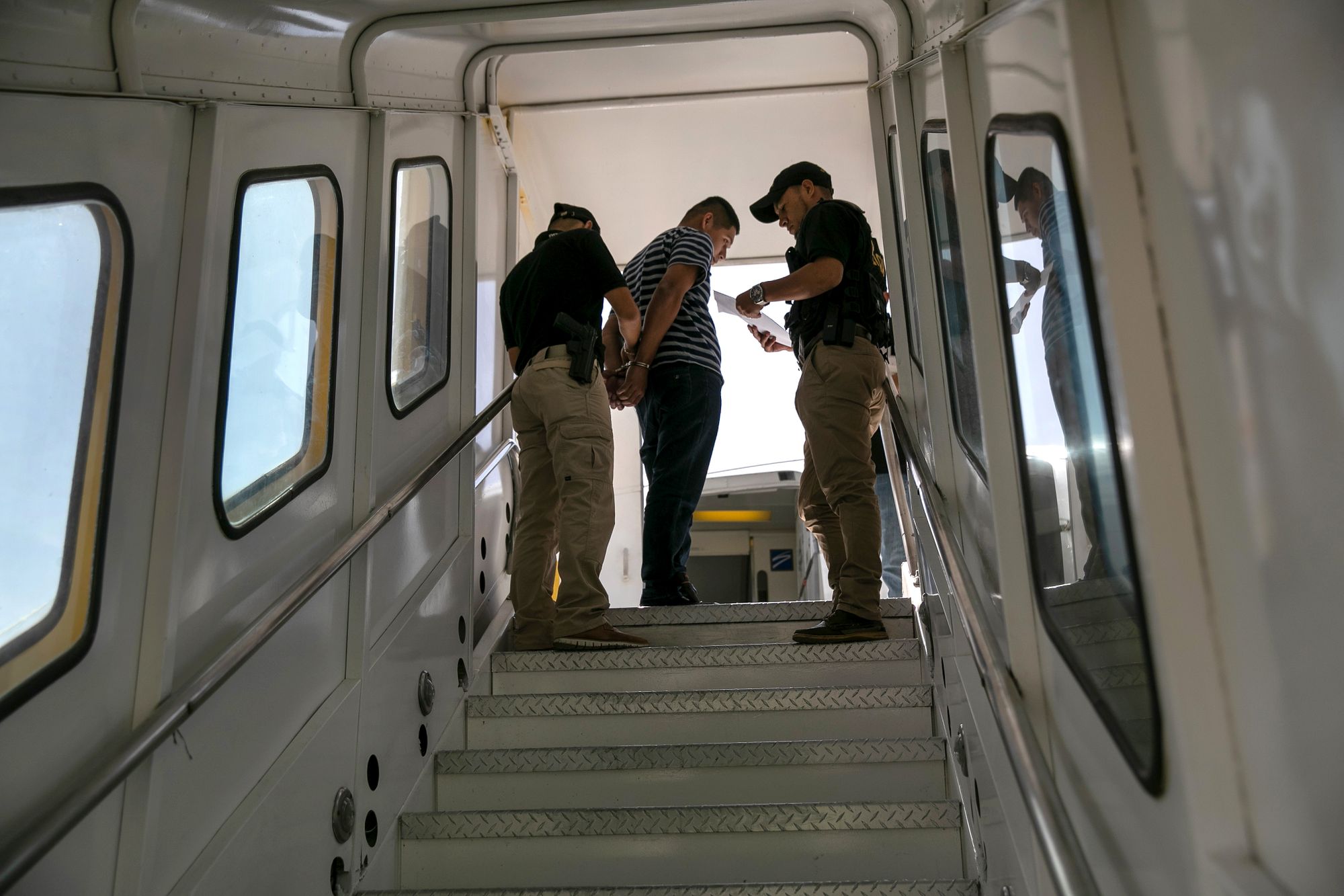 A man looks down with his arms behind his back, as he is being handcuffed by a police officer at the top of a jet bridge walkway. An officer to the right reads from a piece of paper. 