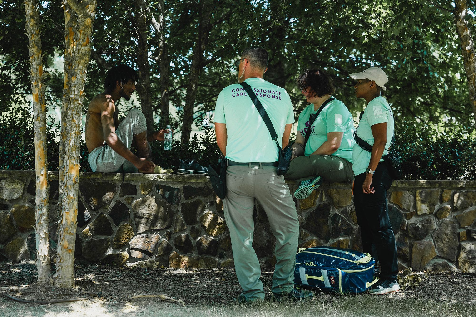 A crisis response team of three people, wearing cargo pants, turquoise shirts and fanny packs, talk to a medium-dark-skinned man sitting on a concrete wall. The man, wearing gray sweatpants, holds a water bottle in one hand.