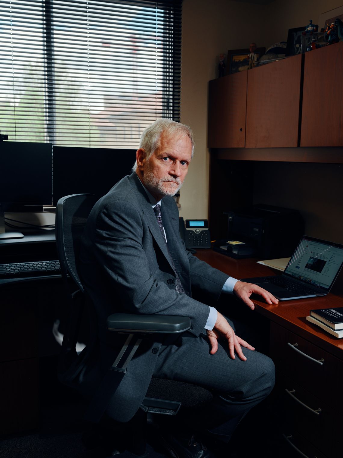 A photo shows a White man with white hair and a beard, wearing a charcoal-colored suit. He is seated in an office facing to the side and looking at the camera.