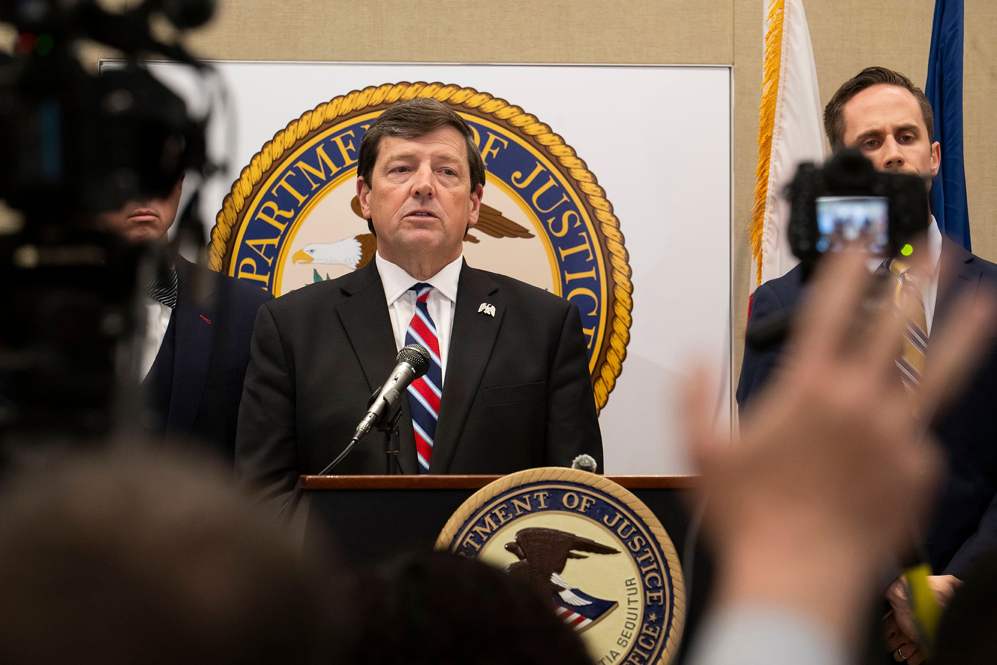 A White man, wearing a black suit and a blue and red tie, talks into a mic while standing at a podium with a U.S. Department of Justice seal on it. A group of reporters holding cameras and microphones stand in front of him. 