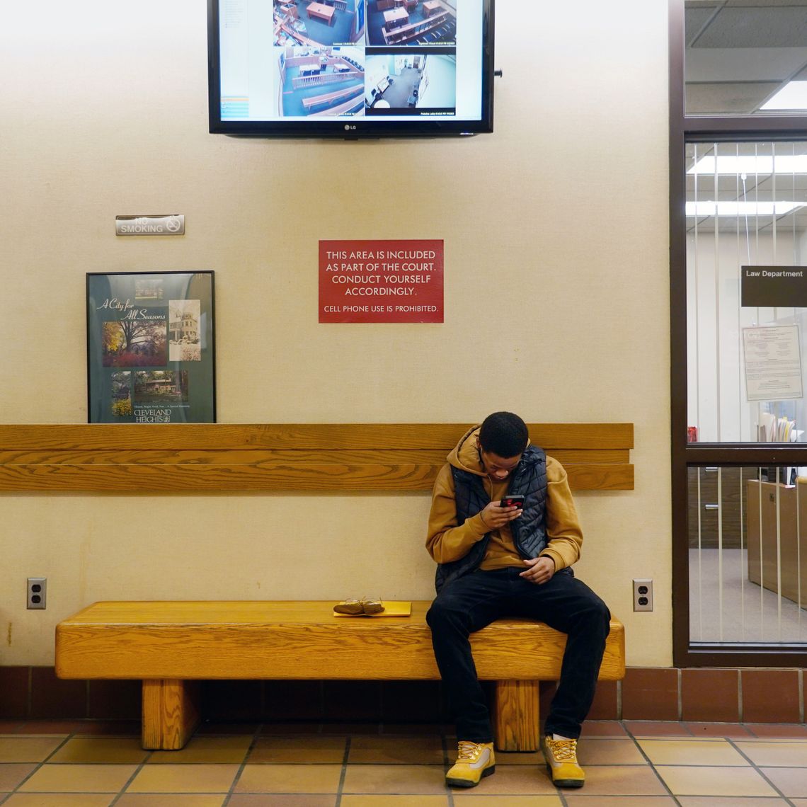 A Black man wearing a dark yellow hoodie and black vest sits on a bench in a court hallway and looks at his phone. 