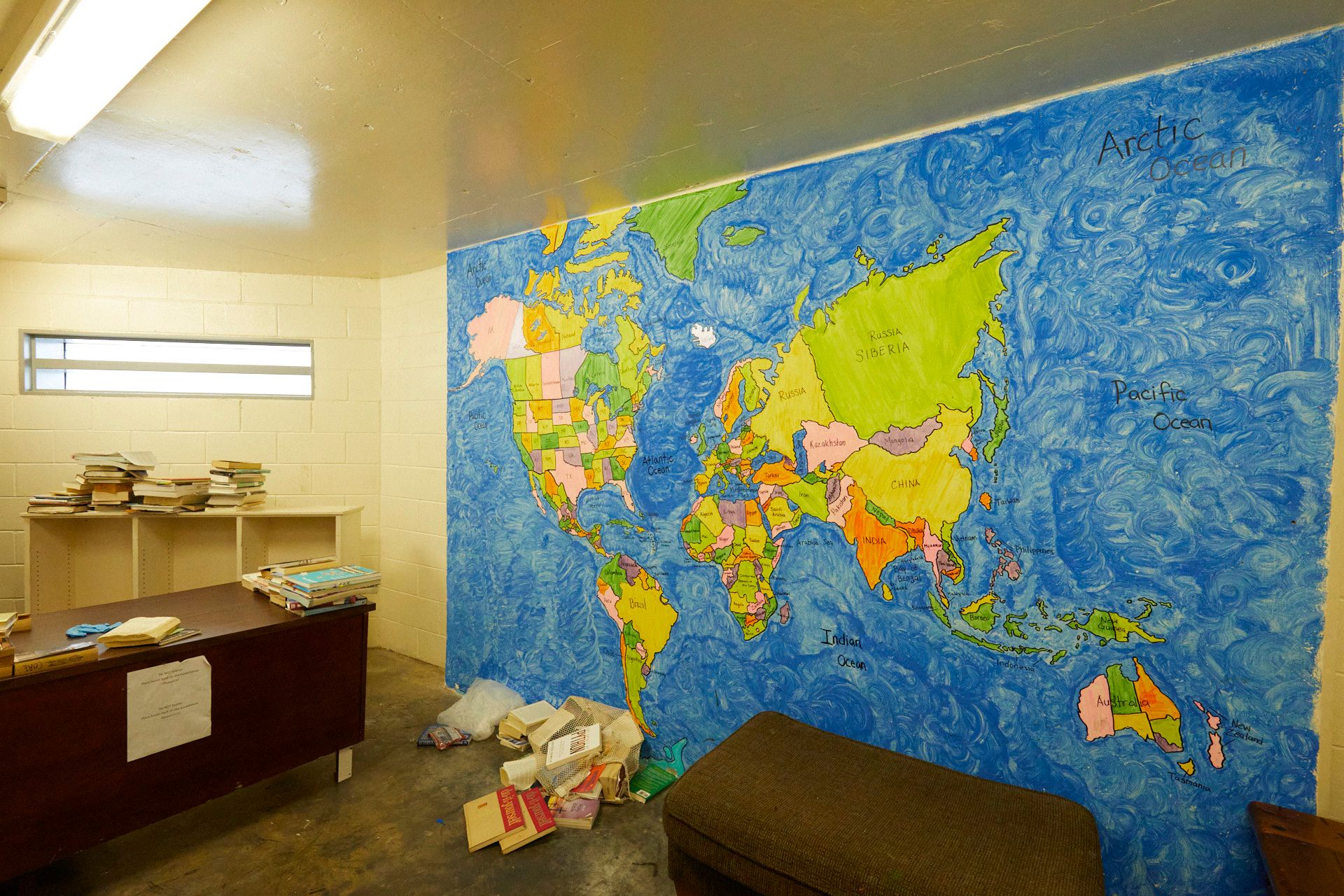 A room shows a painted map of the world, with blue for the ocean, and shades of green, pink, orange and purple for the countries and states. A desk to the left has books piled on it. 