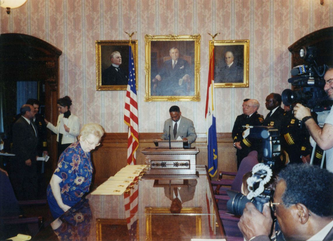 A Black official stands at a podium in a paneled room with portraits of White men on the wall and two flags. He is being photographed by members of the press on the side. An elderly woman in a floral shirt moves past a table with documents.