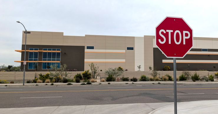 A photo shows a brown and tan warehouse, a road and a stop sign. 