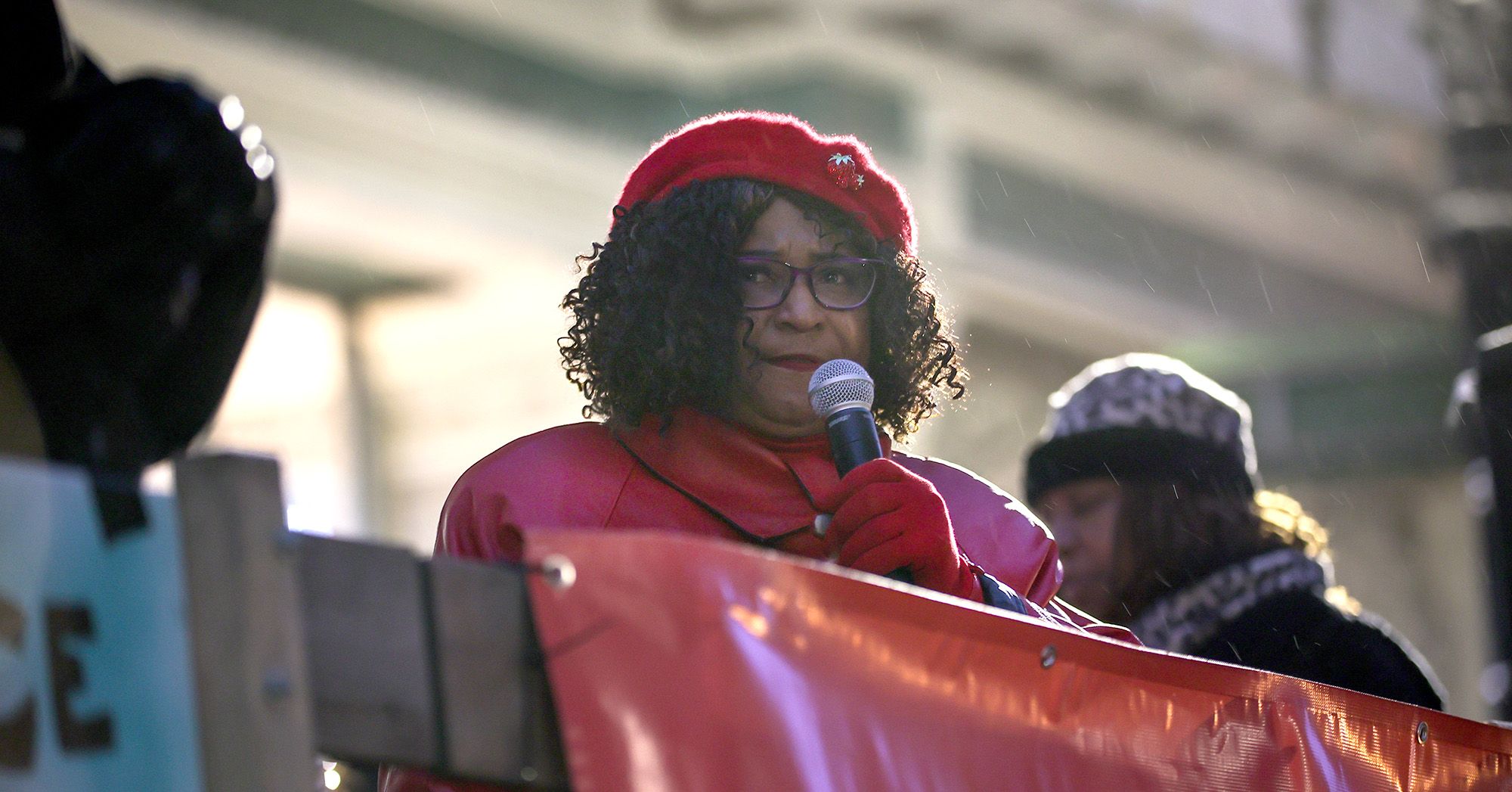 A Black female attorney wearing a red coat, hat and gloves, speaks into a mic during a protest. 