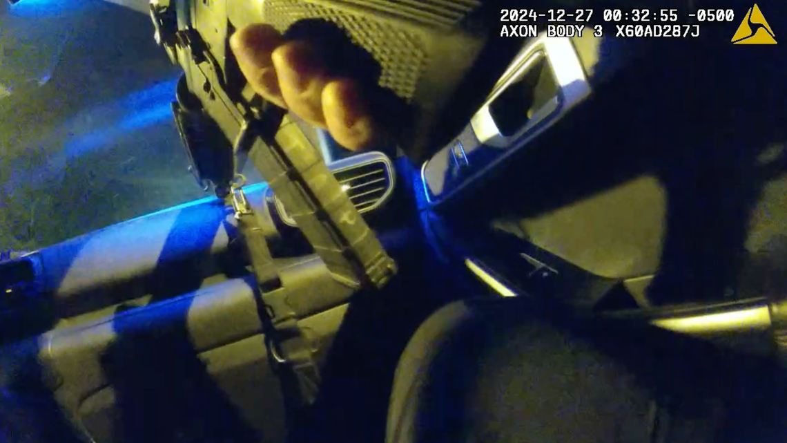 A screenshot of bodycam footage shows a person’s hand holding an assault rifle inside a vehicle at night.