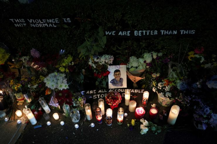 A photo shows a drawing of a man with medium skin tone smiling. The drawing is placed in a memorial with flowers and lit candles. There are posters hanging above the memorial that read, “This violence is not normal,” and, “We are better than this.”