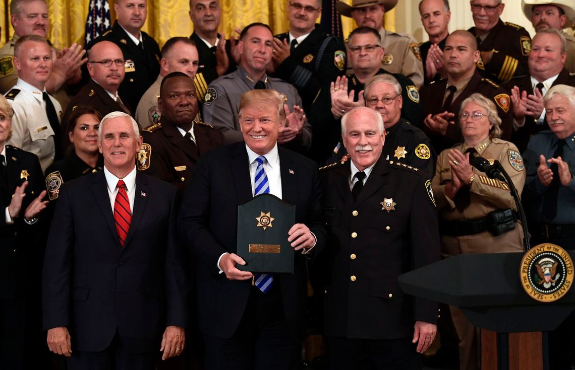 President Donald Trump, a White man, holds a plaque as more than a dozen people stand next to him or behind him at The White House, clapping or smiling. 