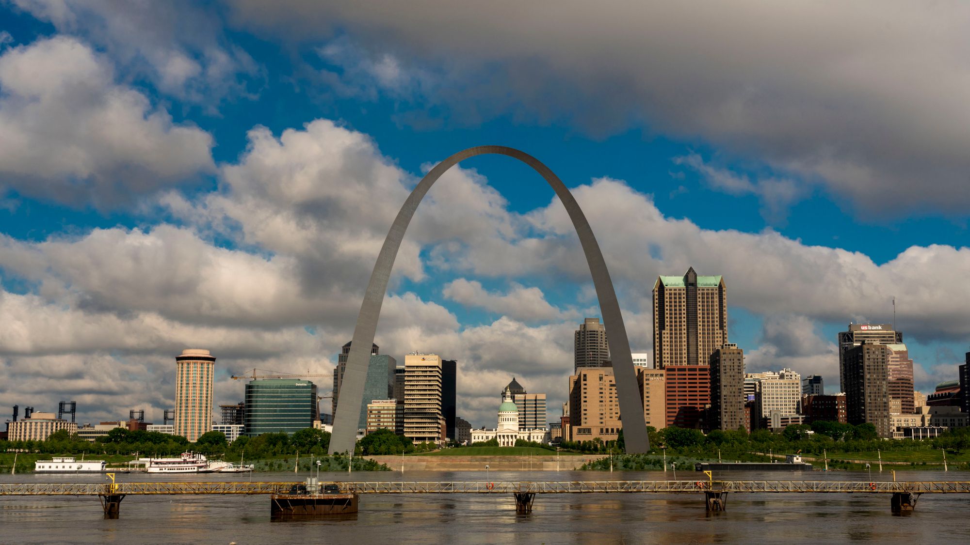 The Mississippi River is visible in the foreground of a photo, with the Gateway Arch in the center.  Buildings are in the background, and a blue sky with clouds is visible. 