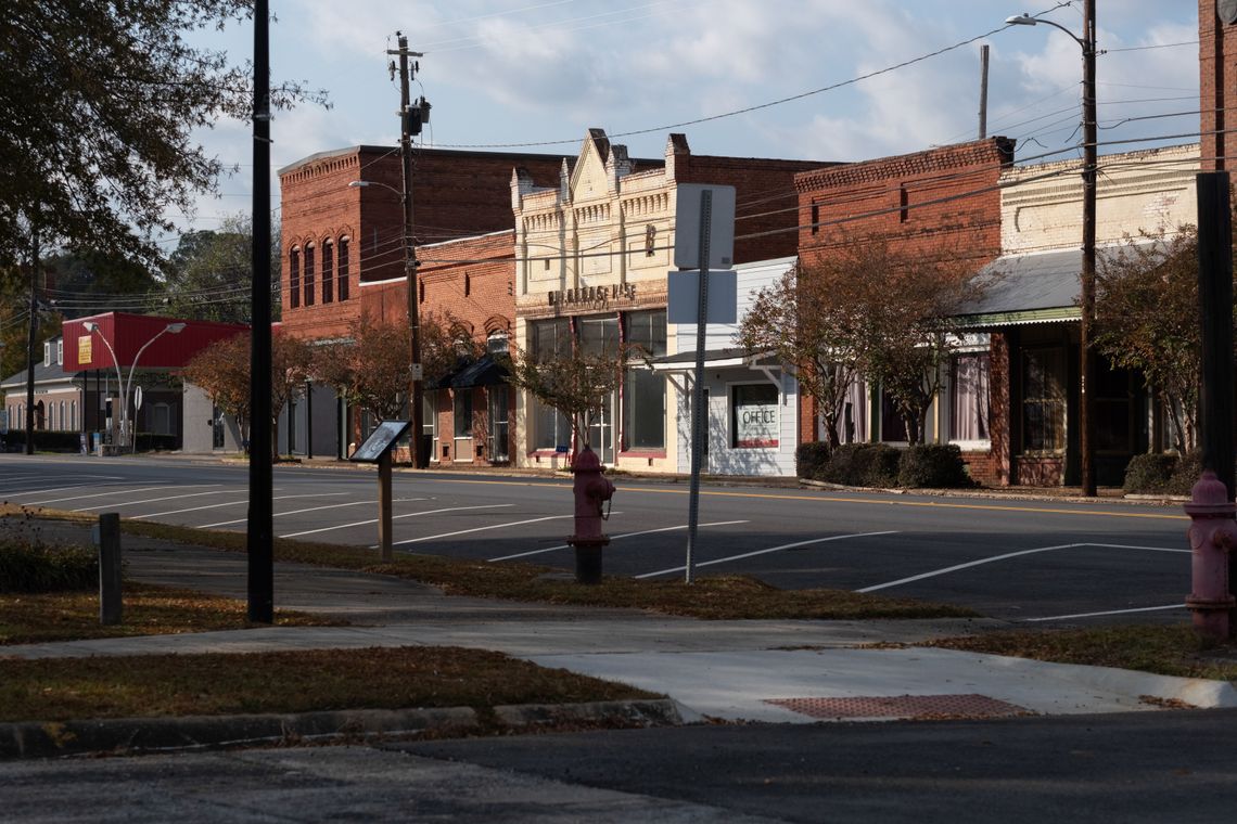 A view of Lumpkin's downtown, which is composed of small brick buildings, including three red ones, two white ones and a pale blue one. All of the parking spaces in front of the buildings are empty.