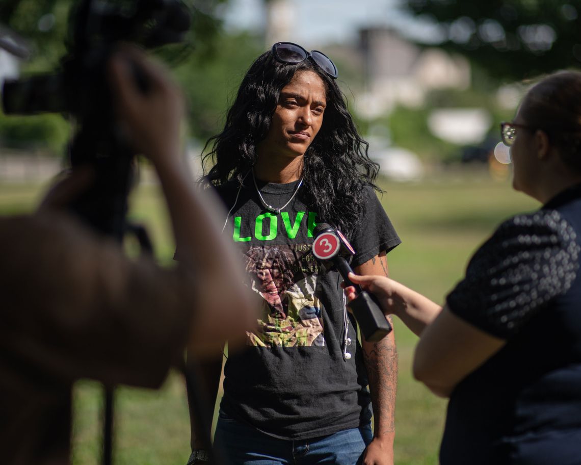 A photo of a young Black woman wearing a black shirt with a photo of her brother and speaking with a reporter holding up a microphone.