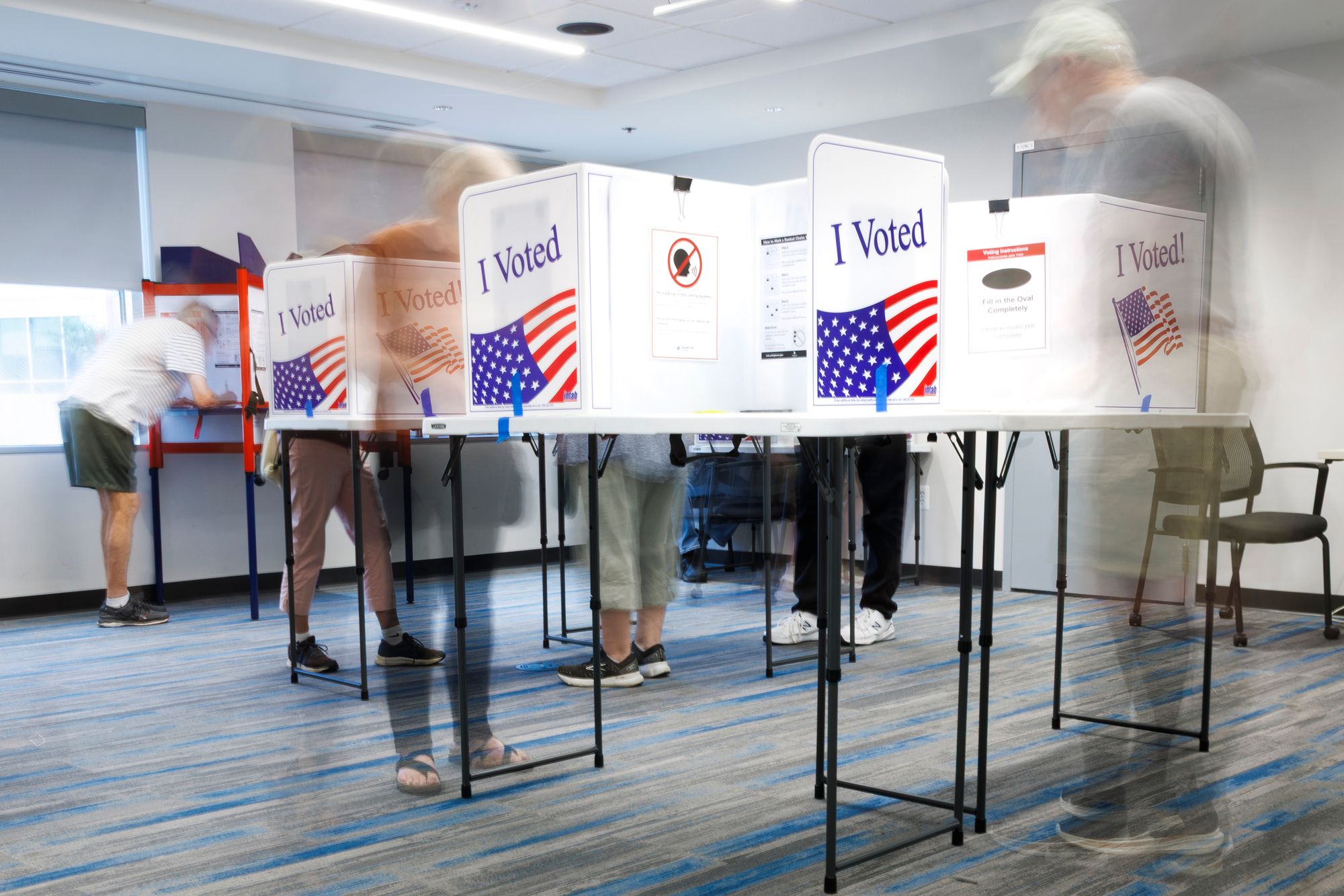 A photo shows people voting in voting booths with the American flag printed on them. The camera used a long exposure so the people look blurred from their movement.