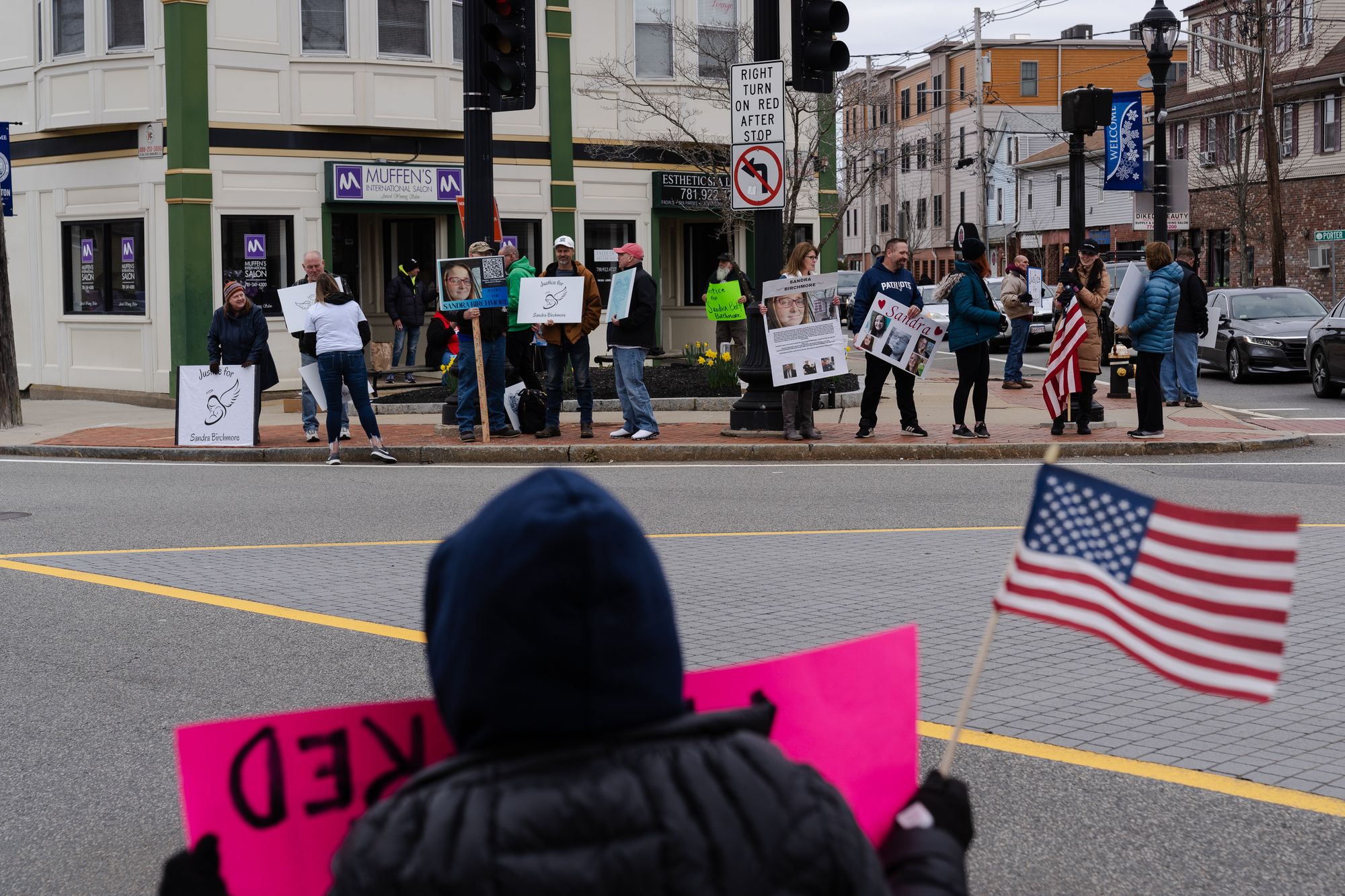 A group of protesters hold signs, some showing photos of Sandra Birchmore, at a street corner.  A person in the foreground is holding a pink sign and a small American flag.  