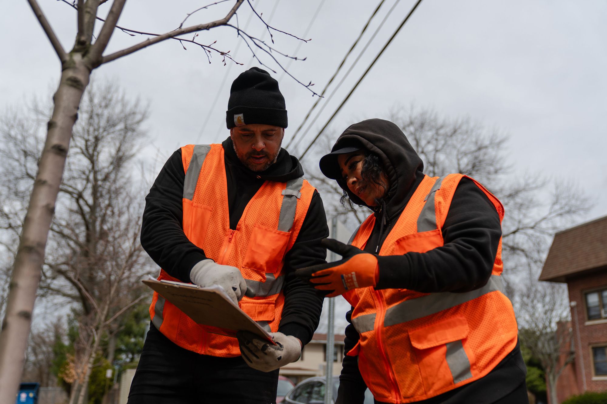 A photo of a man and a woman with medium skin tones looking at a clipboard while wearing orange safety vests. 