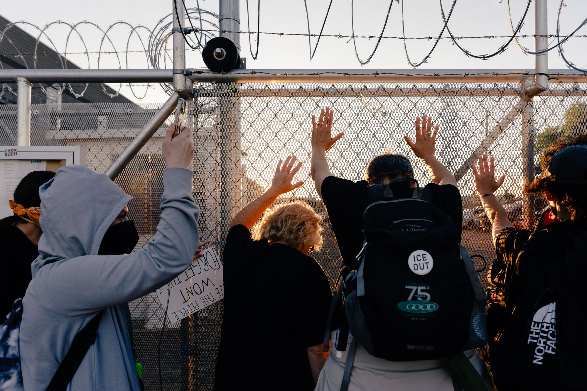 A photo shows a group of people with their hands on a fence with barbed wire at the top. They are outside the facility, facing in. One person has a button on their backpack that says “ICE Out,” and another wears a light-blue hooded jacket and a face mask.  