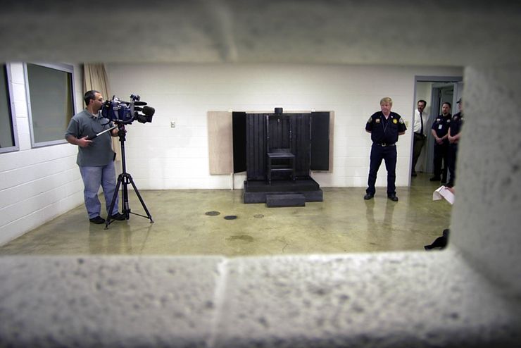 A photo shows a metal chair, backed by two-by-fours with layers of Kevlar attached to the wall. A person operating a video camera is standing on the left side of the room. On the right, three men in dark correctional staff uniforms stand with a man in a white shirt and black tie.  