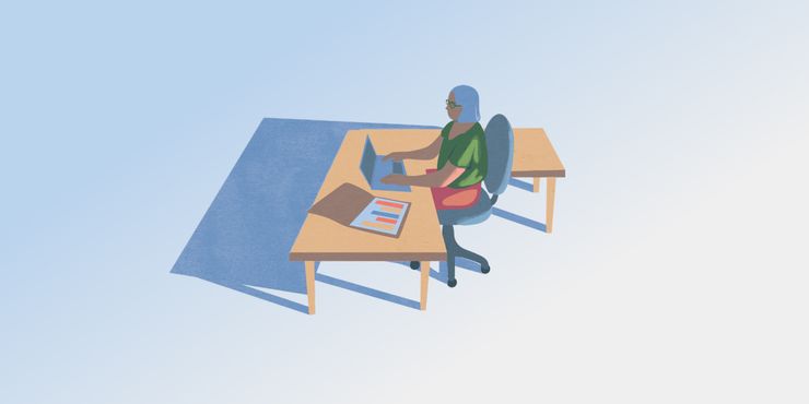 A person with shoulder-length white hair and a green shirt sits at an L-shaped desk typing on a computer and examining charts.