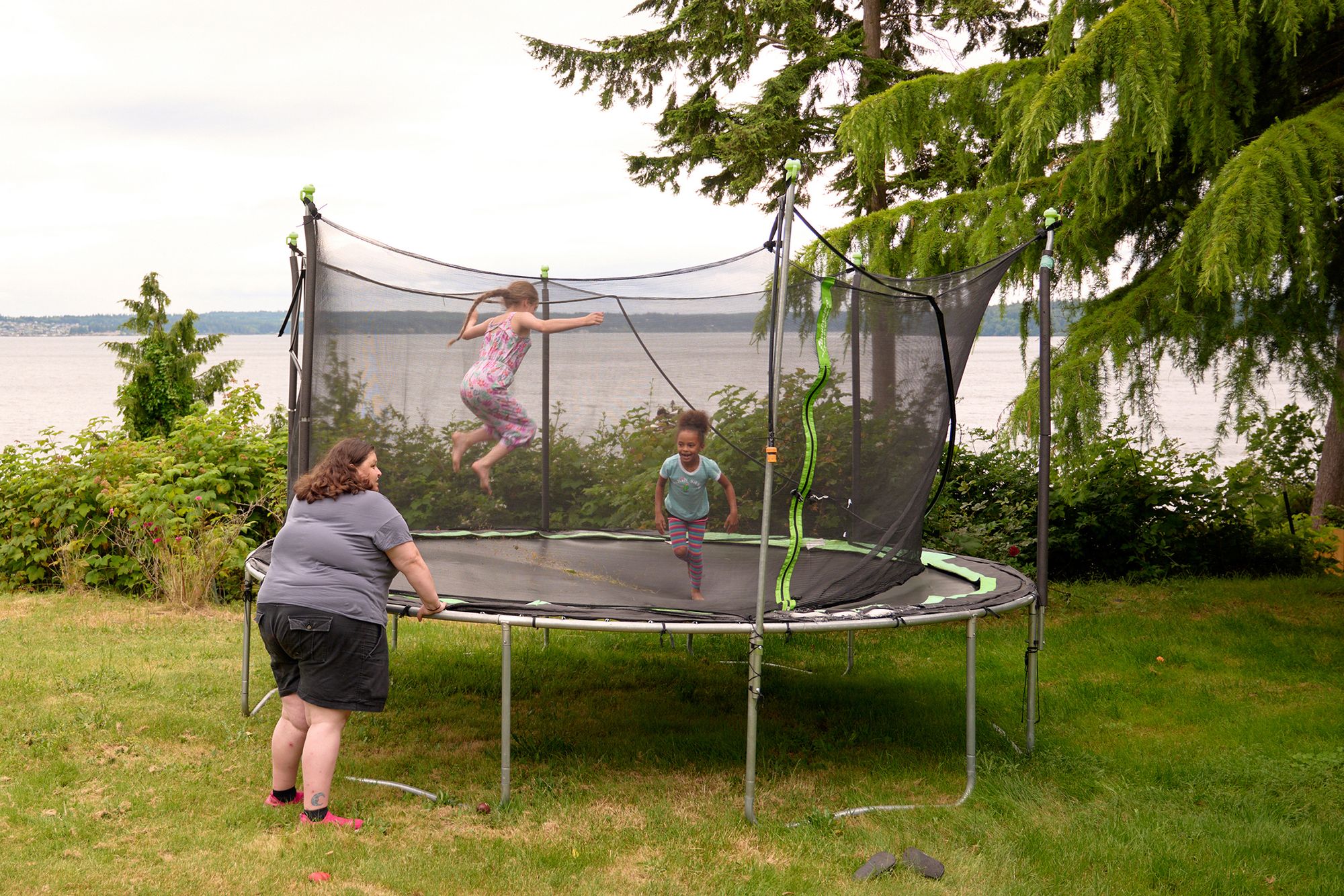 A woman with light-toned skin and a gray T-shirt watches as a young girl with light-toned skin and a young girl with medium-toned skin jump on an outdoor trampoline. 