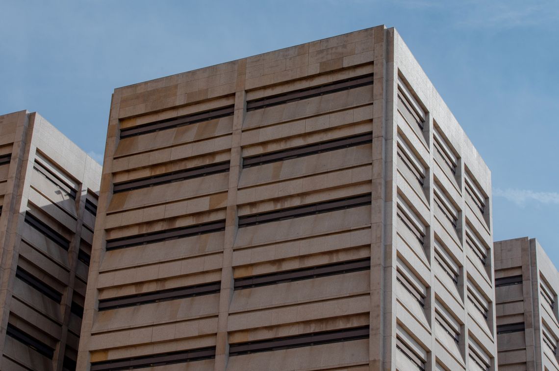 A photo shows a multistory building complex with narrow, horizontal windows. One side of the building is in shade while the other side is in harsh sunlight. Blue sky is visible above the building complex.