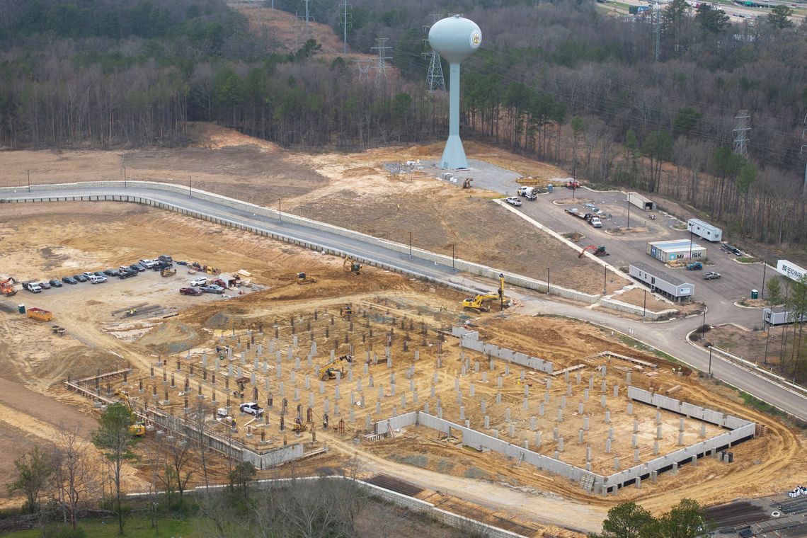 An aerial photo shows a site in the early stages of construction. Cars are parked in a lot near the site, which is by a road and water tower.