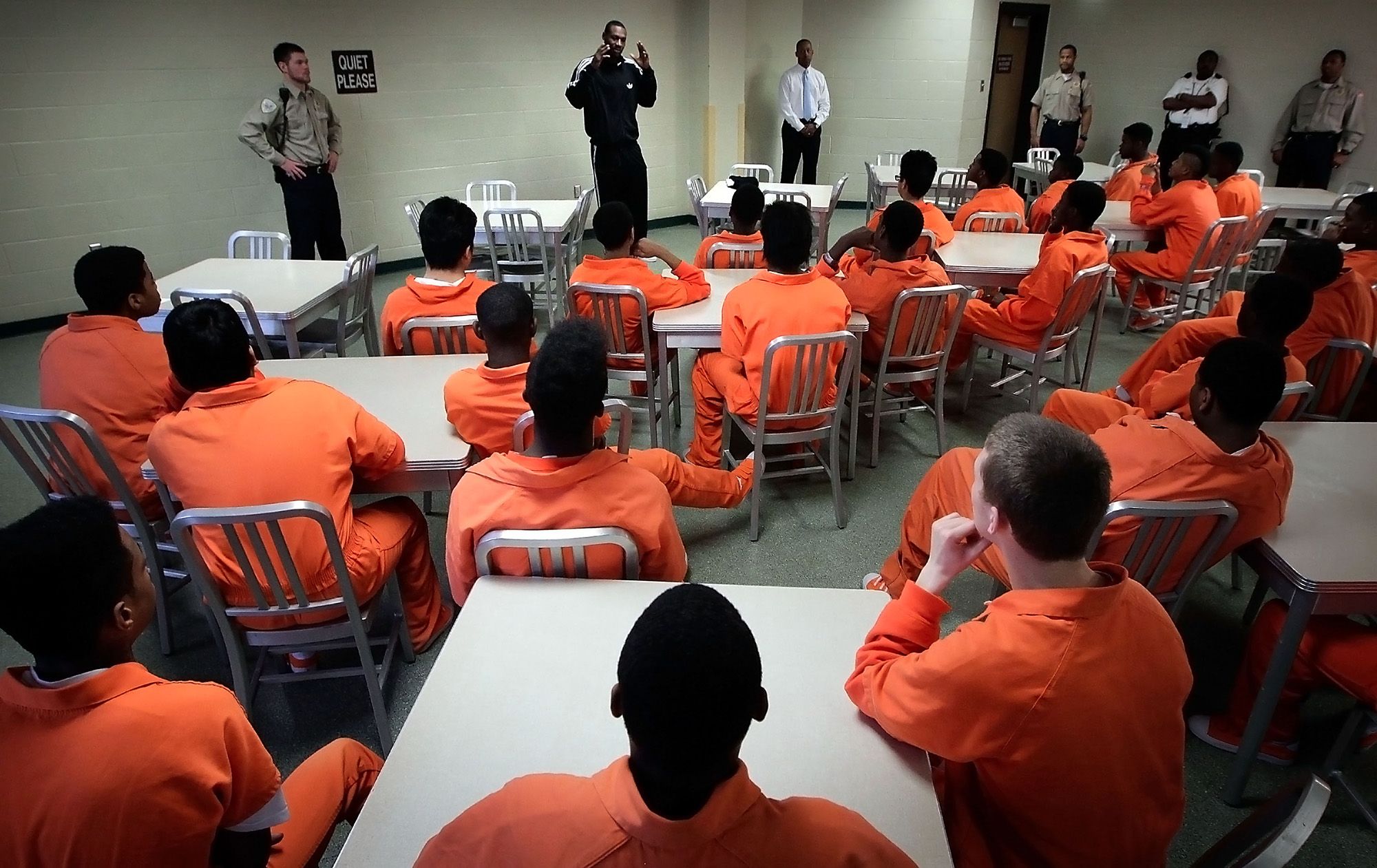 A Black man in a tracksuit stands as he speaks to teenagers, dressed in orange uniforms, sitting inside a room. Officers monitor the teenagers while standing on the side.
