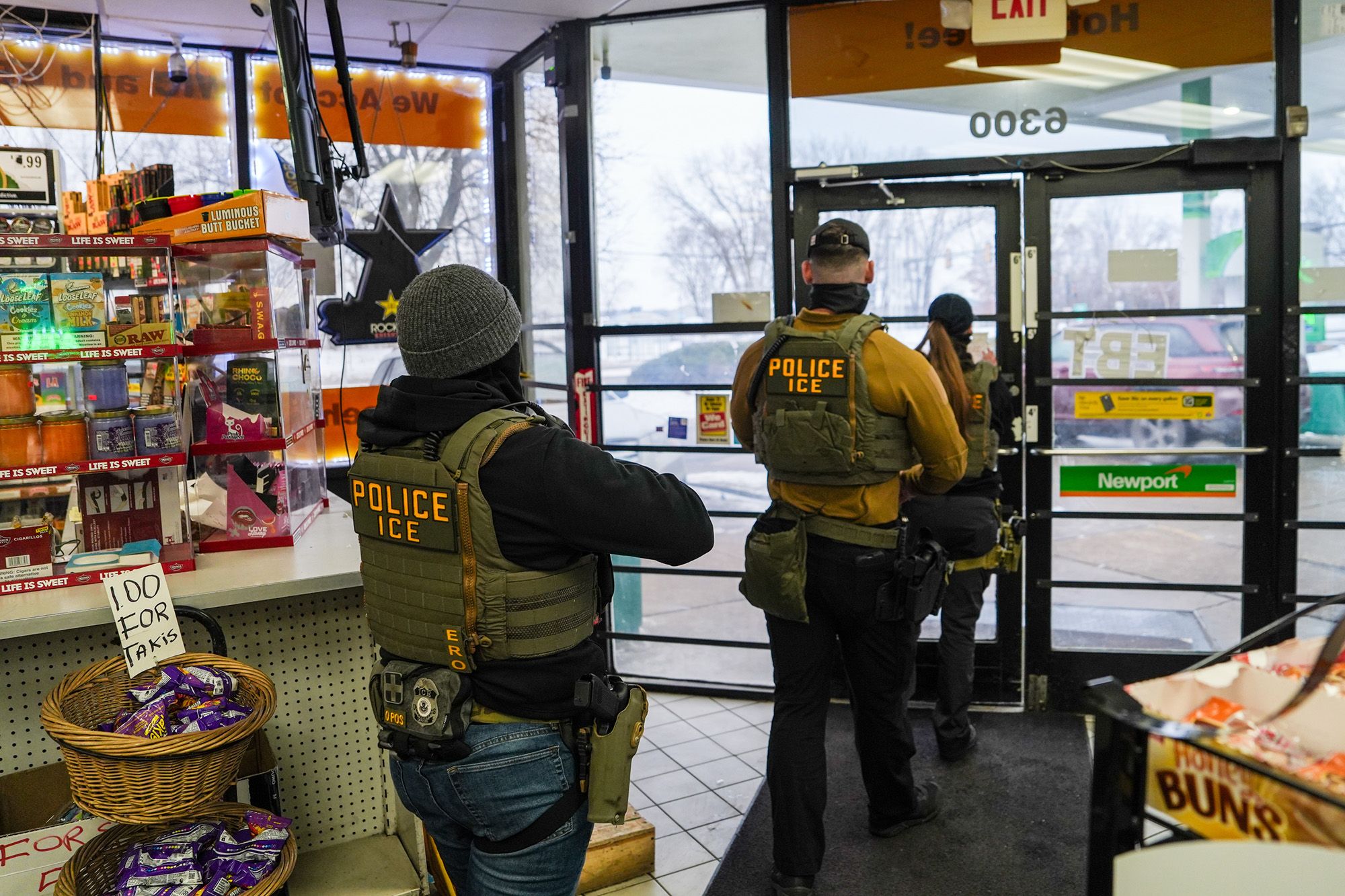 Three Immigration and Customs Enforcement agents, wearing civilian clothes, beanies and vests with labels that read "POLICE ICE," exit out of a gas station.  