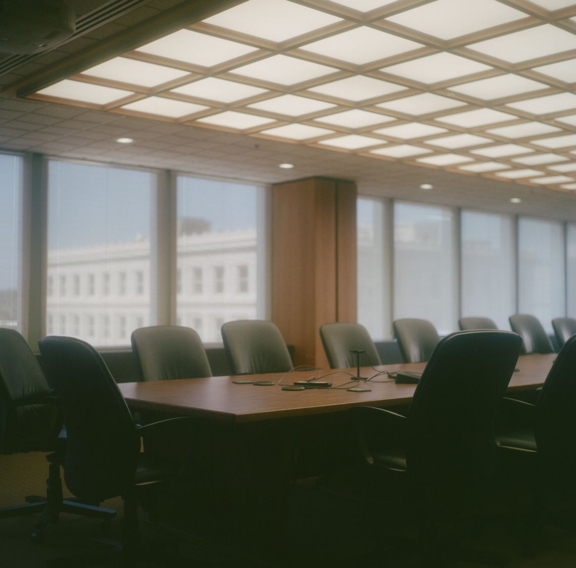 A color photo of a wooden conference table and office chairs in a room with large windows. 