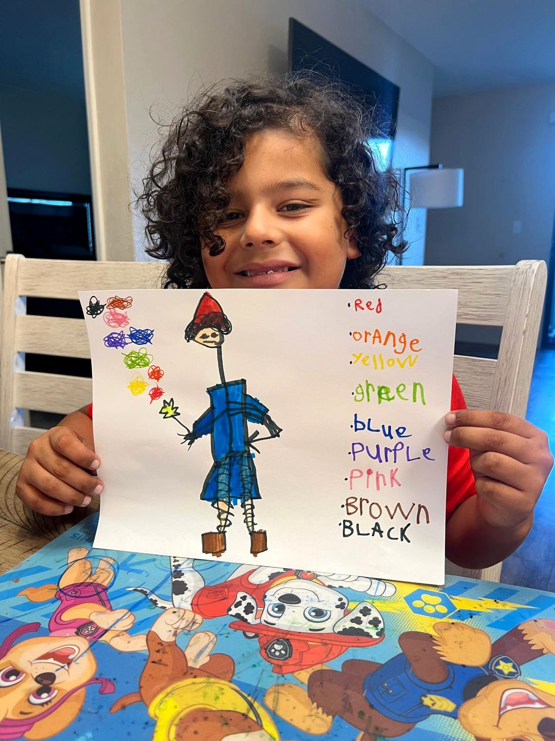 A photo shows a boy with medium-toned skin and curly dark brown hair holding up a colorful drawing of a clothed stick figure, with the words “Red orange yellow green blue purple pink brown black” written in those colors on the right side. He sits at a table that has a tablecloth with a design of cartoon dogs from “Paw Patrol.”