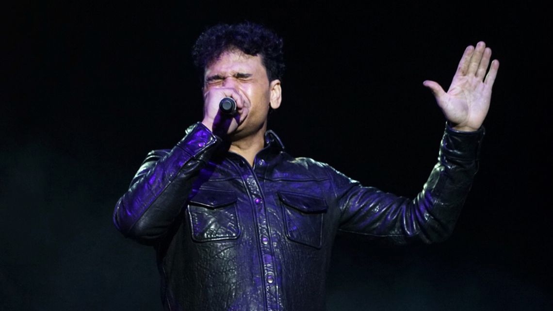 A photo shows a man with medium-toned skin wearing a black leather jacket and singing into a microphone.