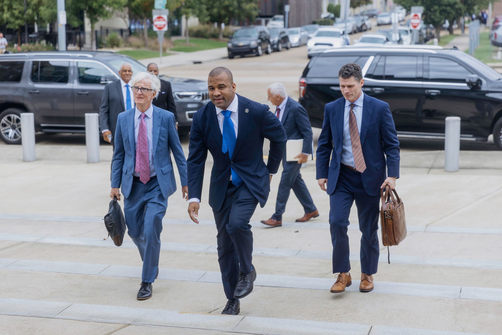 A Black man in a navy suit and blue tie walks up the steps of a courthouse with two White men wearing blue suits and holding leather briefcases.