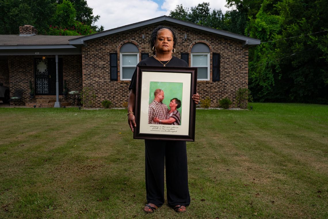 A photo shows a Black woman with curly hair in a ponytail holding a framed picture while posing for a portrait in front of her home. The framed picture shows her embracing and looking toward her son, a Black man with a plaid shirt and glasses, in front of a green background.