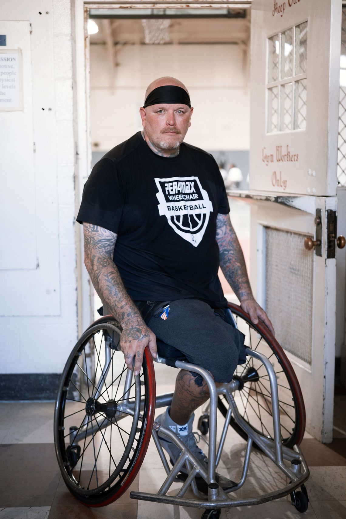 A photo shows a White man wearing a black T-shirt in a wheelchair, wearing a sweatband around his head. He has tattoos on both his arms. 