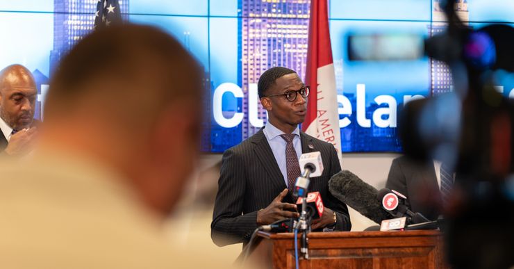 A Black man with short hair in a gray suit stands at a podium during a press conference. 