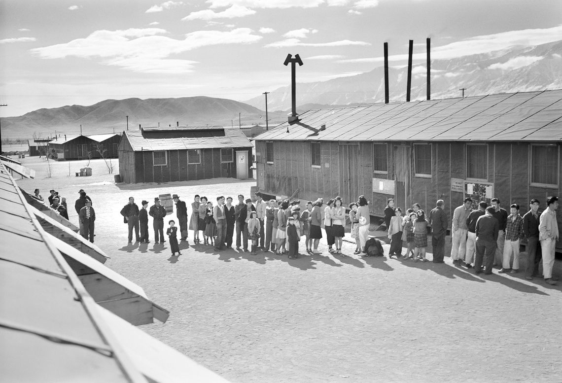 A black-and-white 1940s photo shows Japanese Americans standing in line outside of a building at the Manzanar Relocation Center in California. 