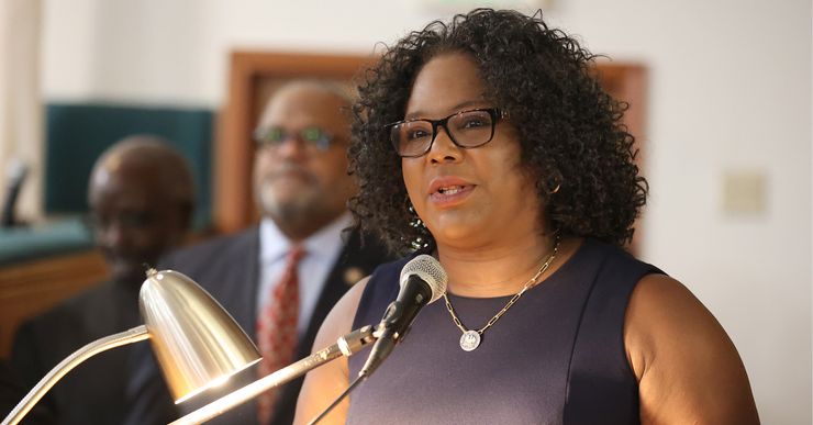 Louisiana Rep. Delisha Boyd, a Black woman wearing glasses and a black dress, speaks into a mic while standing in front of a podium. Two Black men wearing suits stand in the background. 