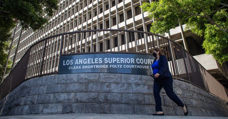 A woman, wearing a KN95 mask, black cardigan, blue blouse, and black pants, walks in front of a sign that reads "Los Angeles Superior Court Clara Shortridge Foltz Courthouse." 