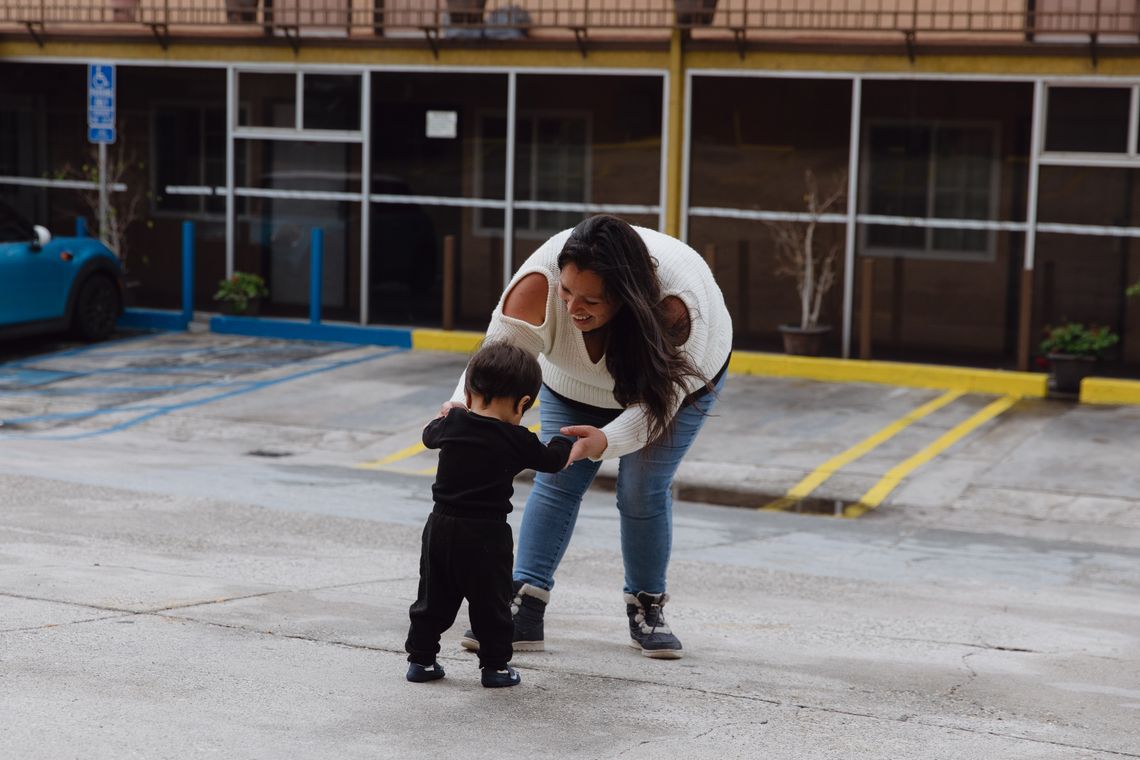 A photo shows Mapes, a Native American woman with long dark hair wearing a white sweater, smile as she holds the hands of her son, an infant with short dark hair and wearing a black shirt and black pants, as he attempts to walk. A motel is visible in the background. 