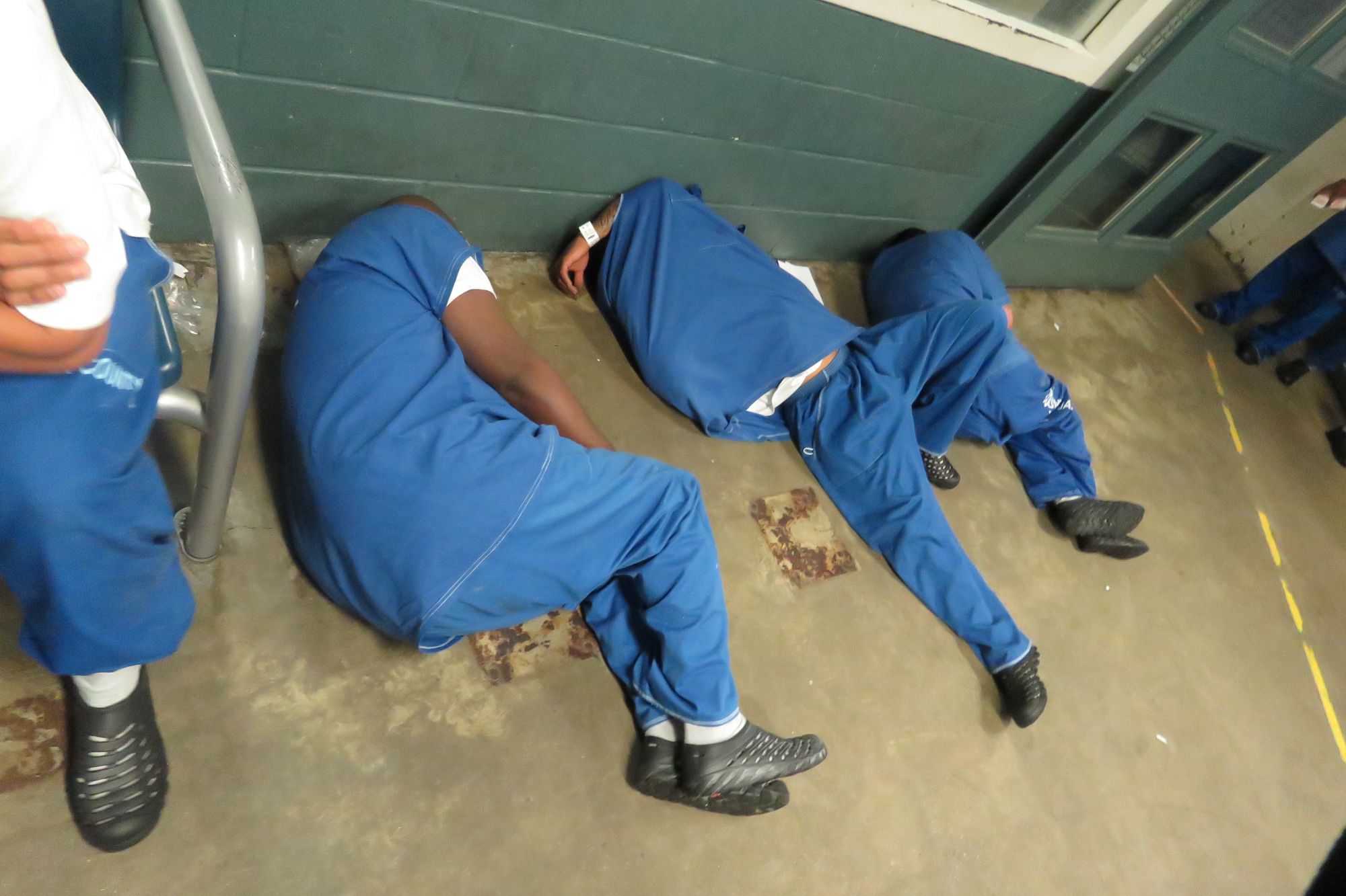 Three men in blue uniforms sleep on the floor, as one sits on a chair nearby.  