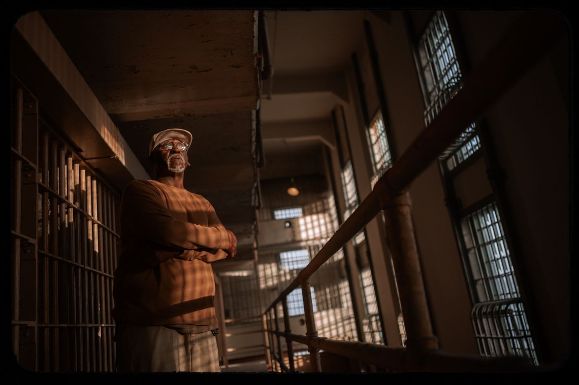 A photo shows Rubin “Jitu” Williams, a Black man wearing glasses, a beige-colored cap and a dark orange long-sleeved shirt, standing with his arms crossed at Alcatraz. Prison cells are visible behind him to the left of the photo, and windows are visible on the right. 