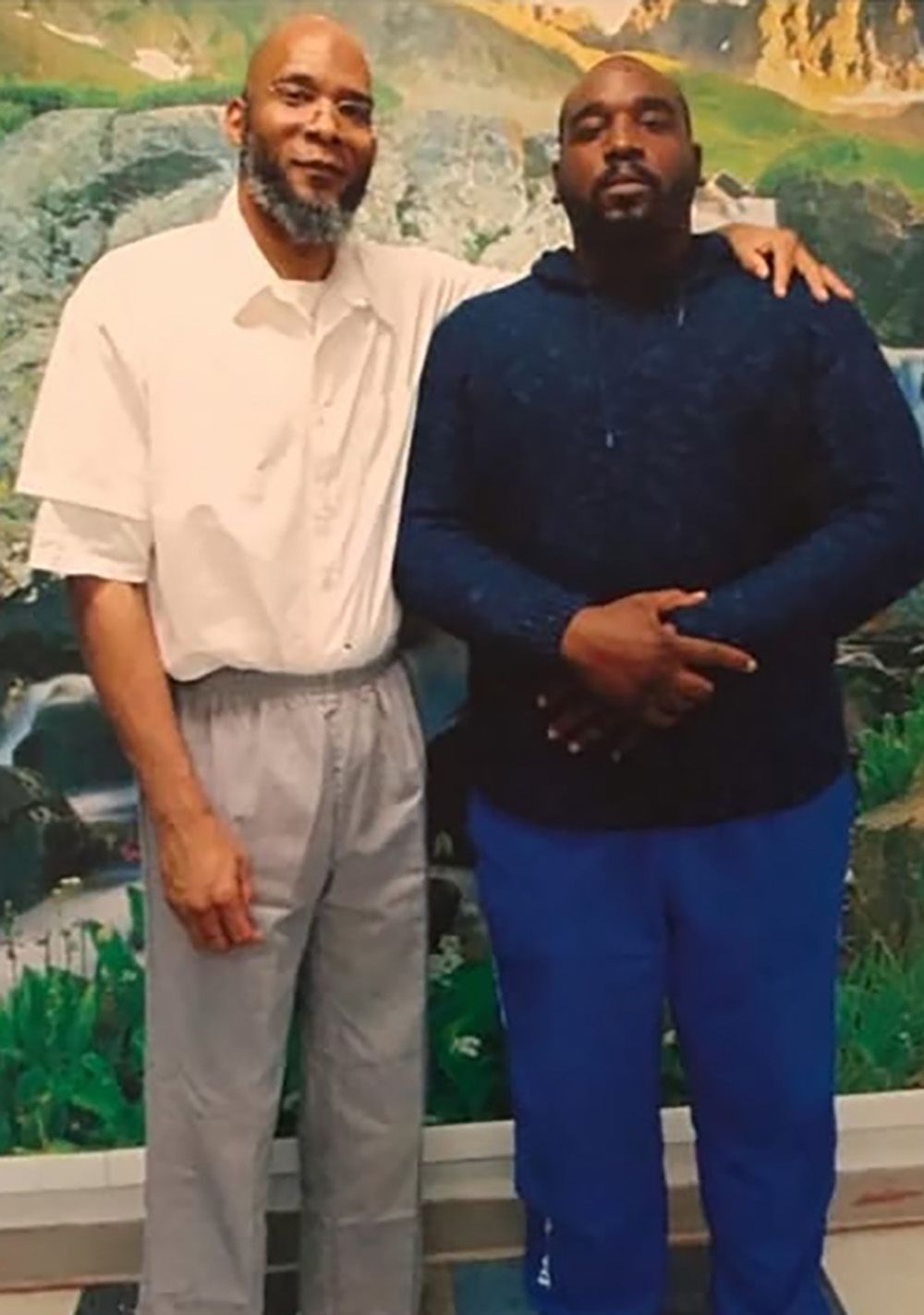 Un hombre Negro con una sudadera y pantalones azules está junto a su padre, posando para una foto, quien viste un uniforme de prisión blanco.