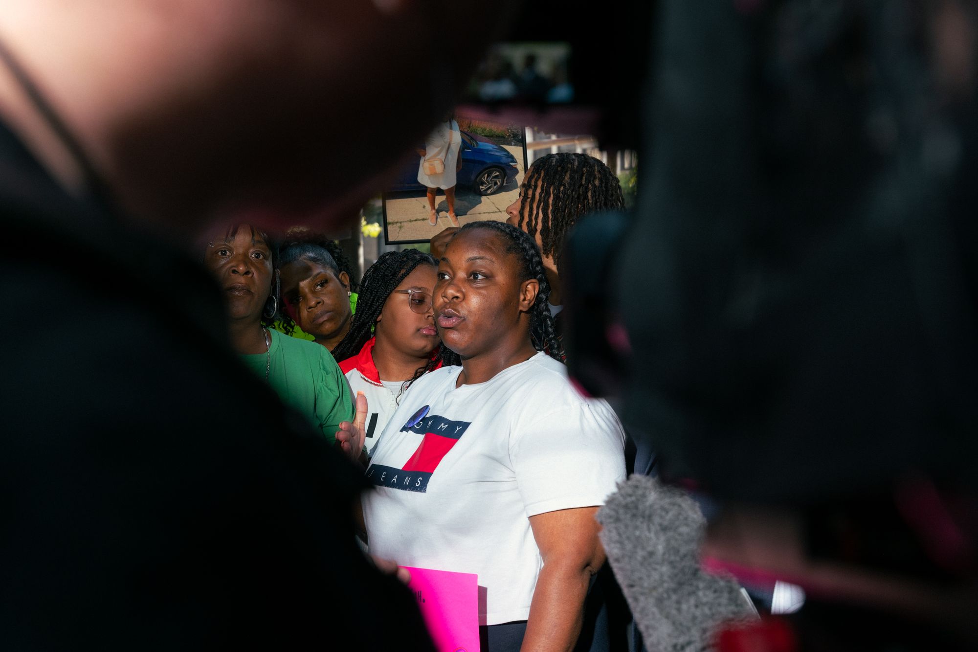 Cearria Elder, a Black woman wearing a white Tommy Hilfiger T-shirt, is visible through the space between a person and a video camera. 