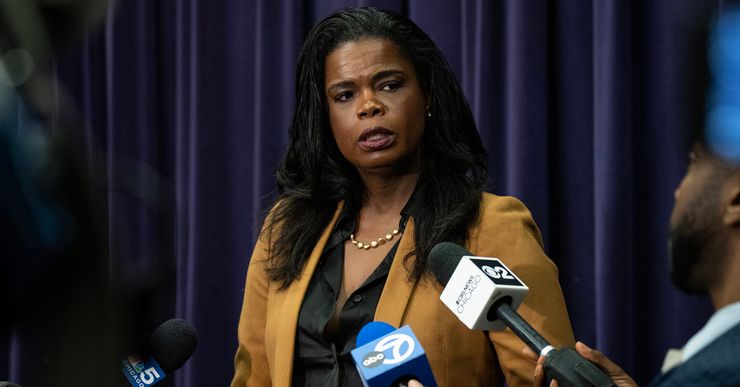 Kim Foxx, a Black woman wearing a tan suit, speaks into mics held by reporters surrounding her. 